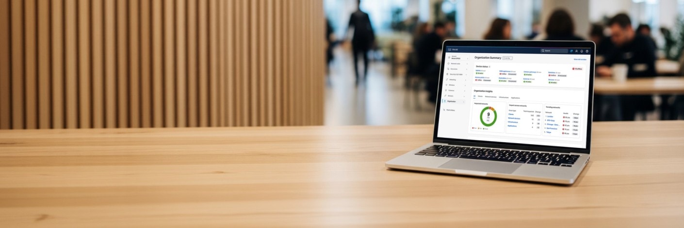 Laptop on a wooden table displaying the Cisco Meraki Dashboard with network metrics, set in a blurred office environment.