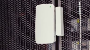 A white wireless sensor mounted on a black mesh server rack door with a green status light.